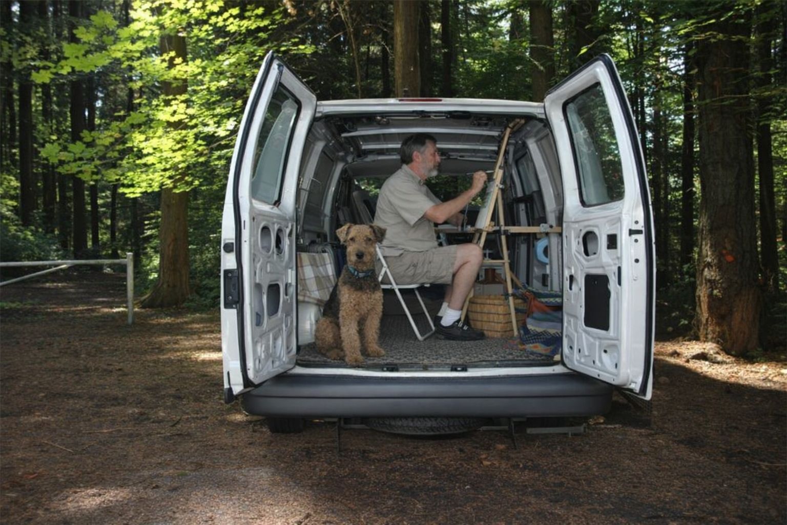 Offener Transporter im Wald: Mann malt an Staffelei, Hund sitzt im Heck